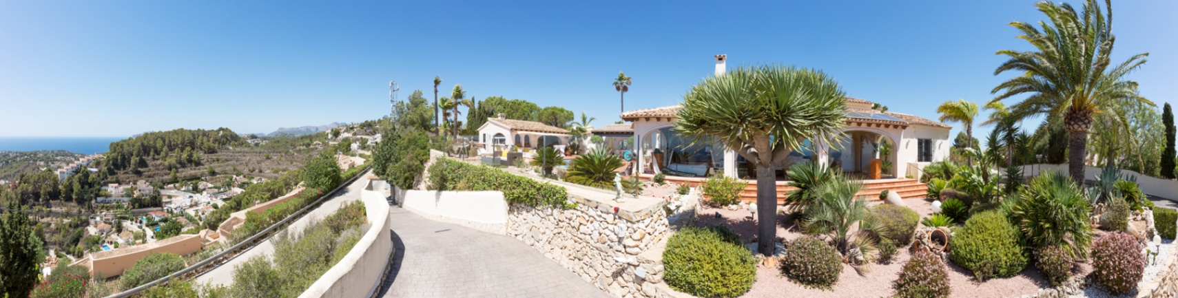 Vue panoramique de l'extérieur de la villa avec une allée pavée, des murs en pierre sèche et un jardin méditerranéen paysager offrant une vue sur la mer.