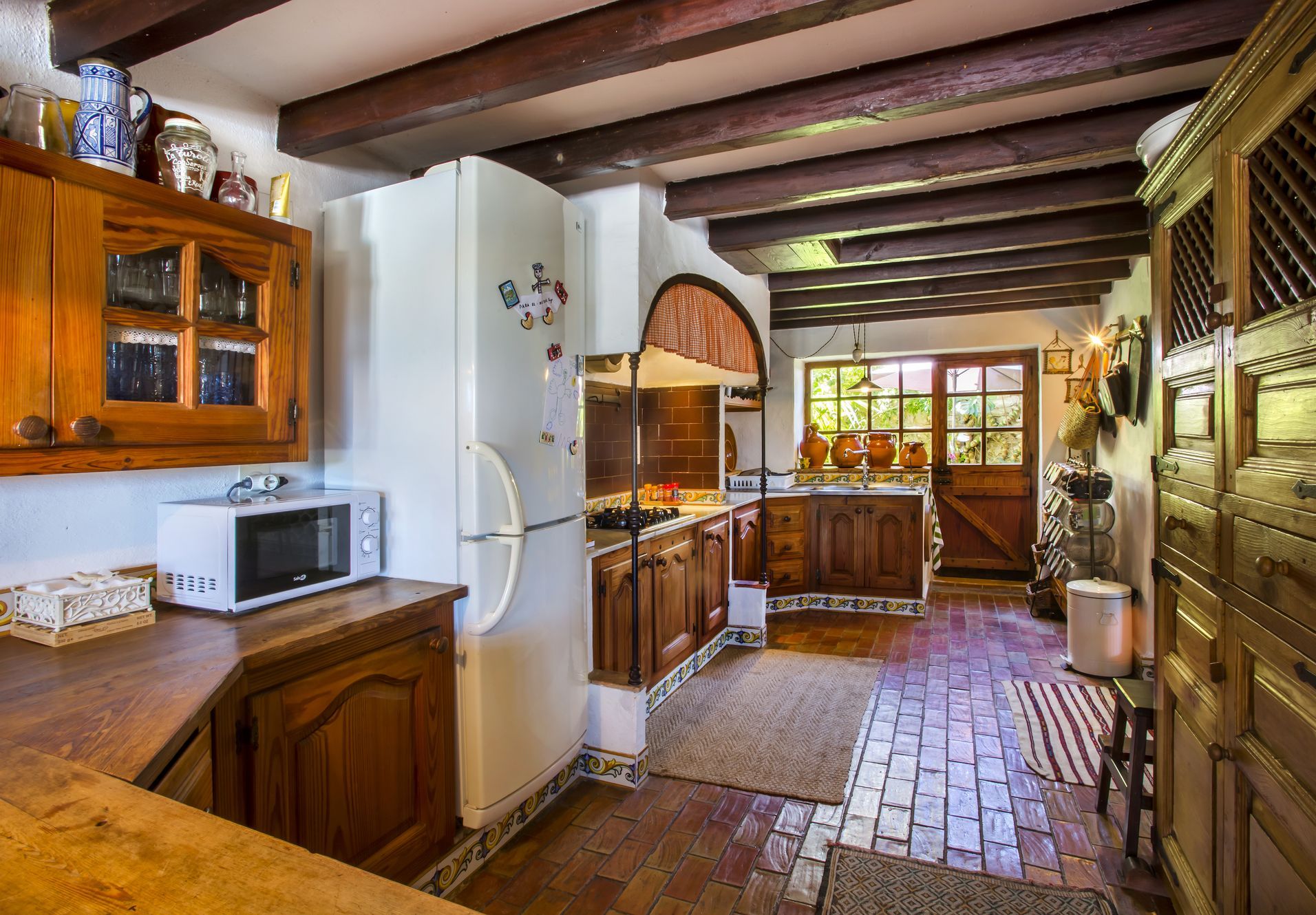 Cuisine rustique avec sols en terre cuite, poutres apparentes et meubles en bois foncé. La lumière naturelle entre par la porte vitrée.