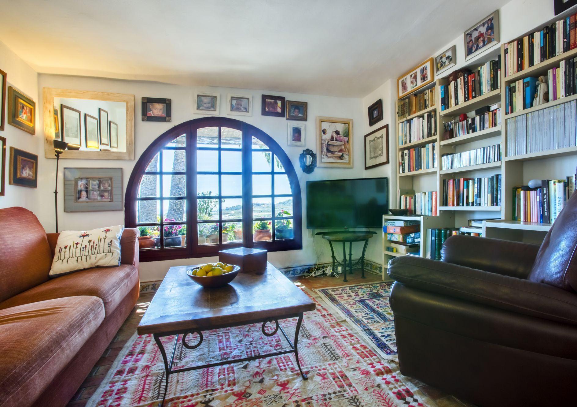 Salon avec fenêtre cintrée et vue, décoré d'un tapis à motifs et d'une bibliothèque remplie de livres.