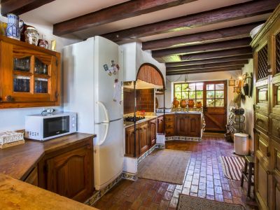 Cuisine rustique avec sols en terre cuite, poutres apparentes et meubles en bois foncé. La lumière naturelle entre par la porte vitrée.