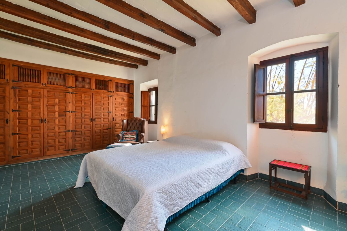 Chambre rustique avec poutres en bois apparentes, placards intégrés en bois sculpté, sol en carrelage vert et fenêtres à volets traditionnels.