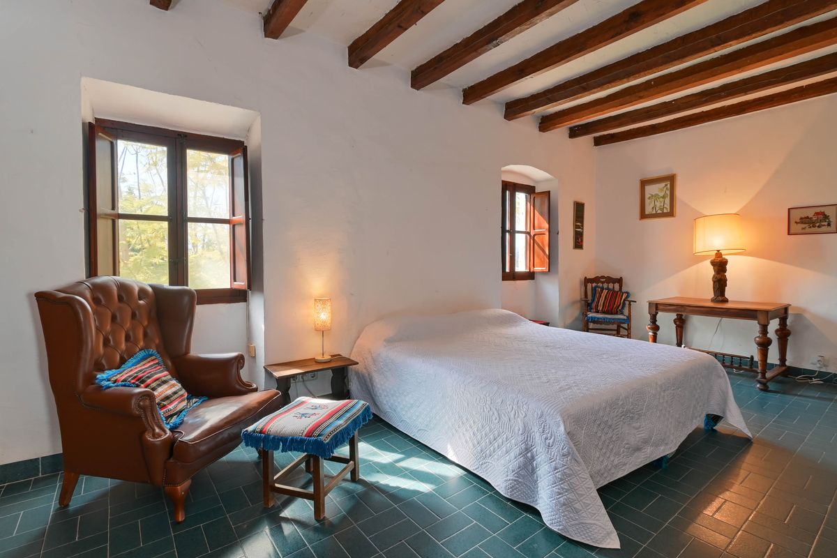 Chambre rustique avec poutres en bois apparentes, sol en carrelage vert et fauteuil à oreilles en cuir près de fenêtres traditionnelles à volets.