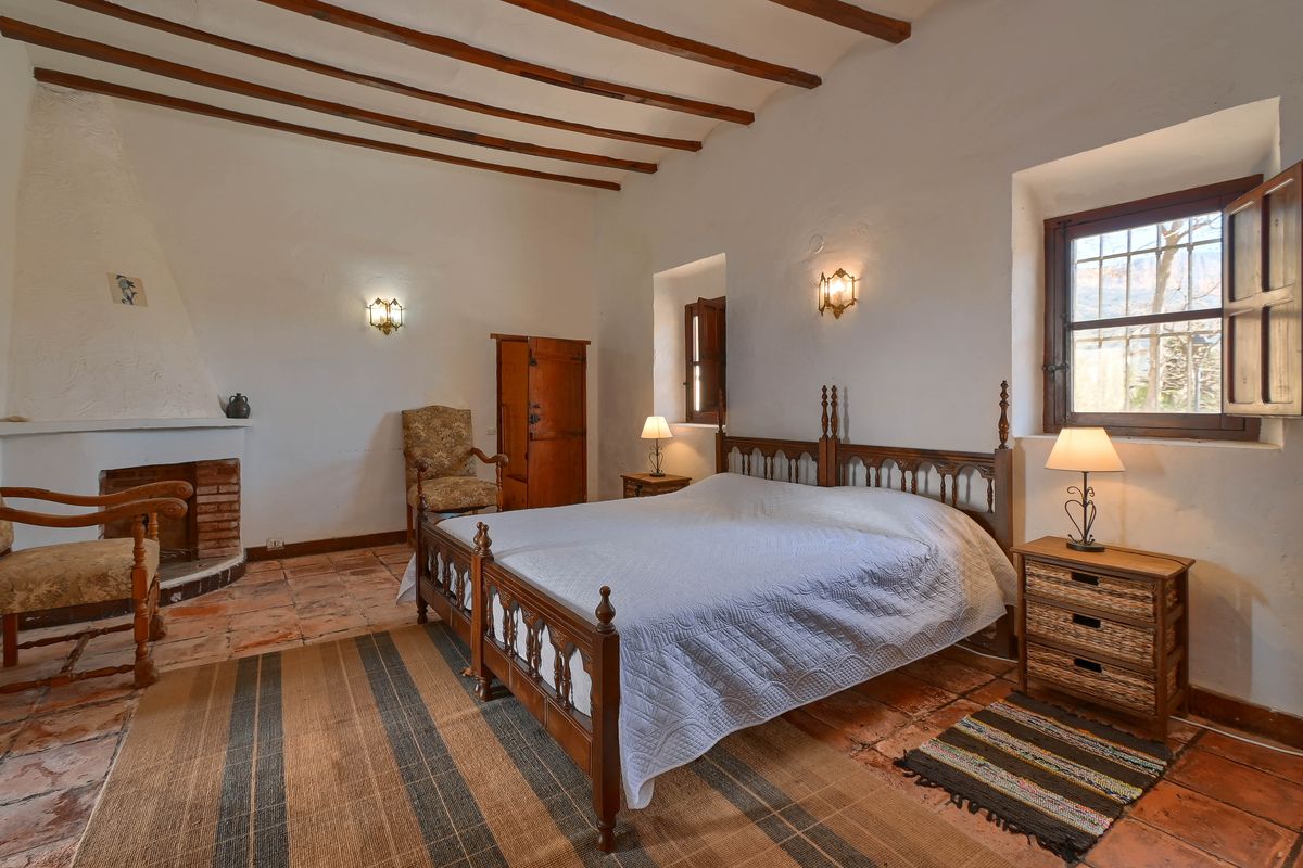 Chambre rustique avec poutres en bois apparentes, sol en terre cuite, cheminée intégrée et fenêtres dotées de volets intérieurs en bois traditionnels.