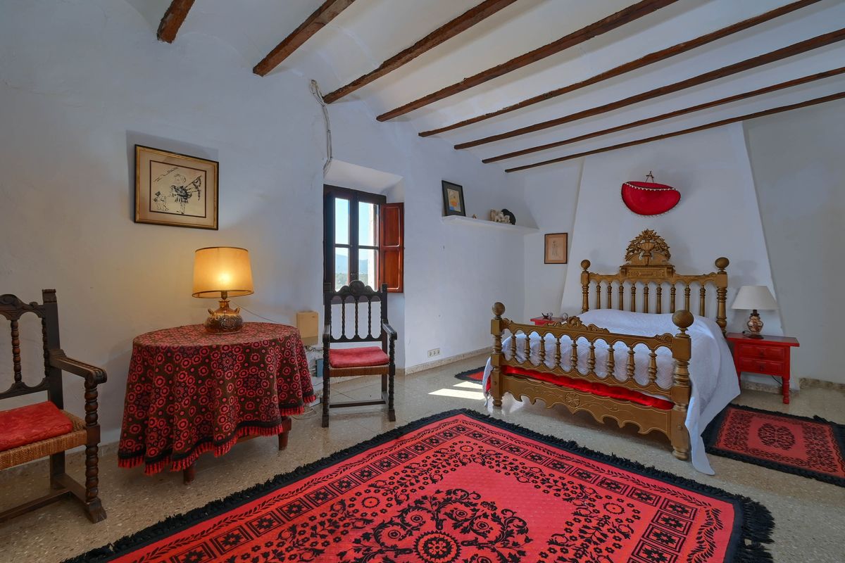 Chambre rustique avec poutres en bois d'origine, cadre de lit sculpté et tapis rouges décoratifs sur un sol en pierre naturelle.