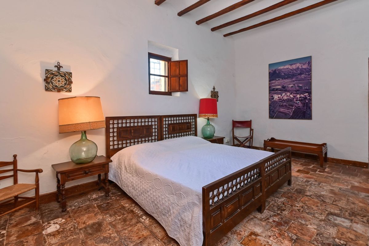 Chambre rustique avec sol en terre cuite, poutres en bois apparentes et lit en bois sculpté. Les lampes en dame-jeanne et les volets traditionnels soulignent le charme authentique.