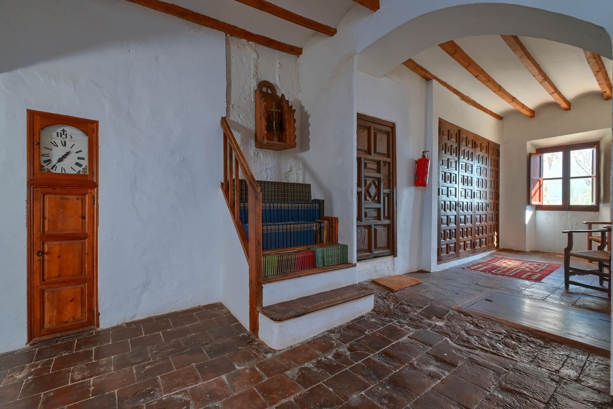 Couloir avec tomettes, poutres apparentes et horloge intégrée. Un petit escalier avec bibliothèque jouxte des portes en bois sculpté de style traditionnel.