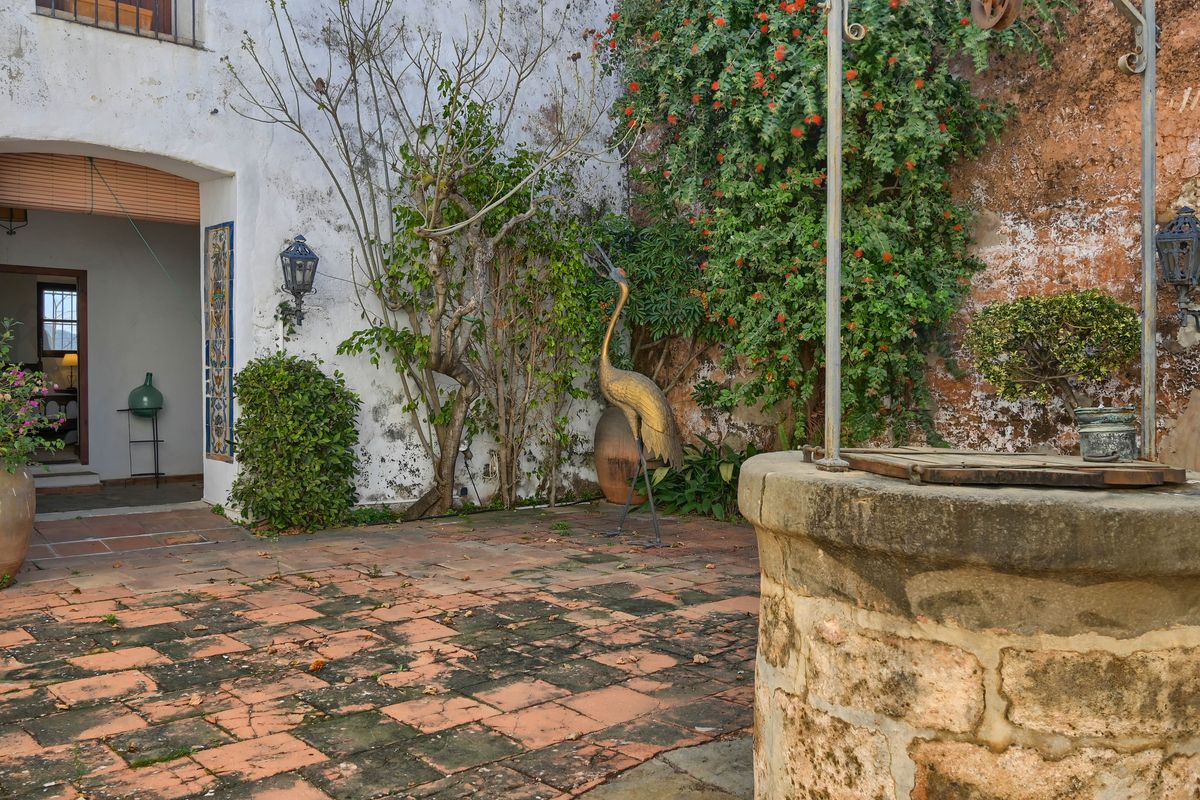 Cour méditerranéenne traditionnelle avec un puits en pierre d'origine, un sol en terre cuite patinée et une sculpture décorative entourée de plantes grimpantes.