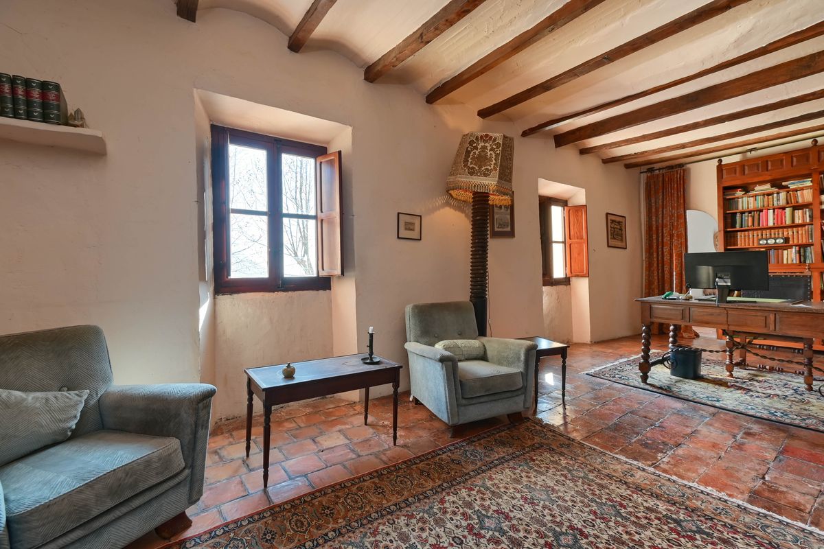 Espace bureau avec poutres en bois apparentes, sol en terre cuite et fenêtres à embrasures profondes. Comprend un bureau ancien et une bibliothèque intégrée.