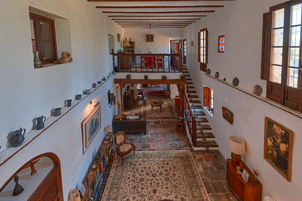 Hall spacieux à double hauteur avec sols en terre cuite, poutres en bois apparentes et un escalier menant à une mezzanine avec balustrade traditionnelle.