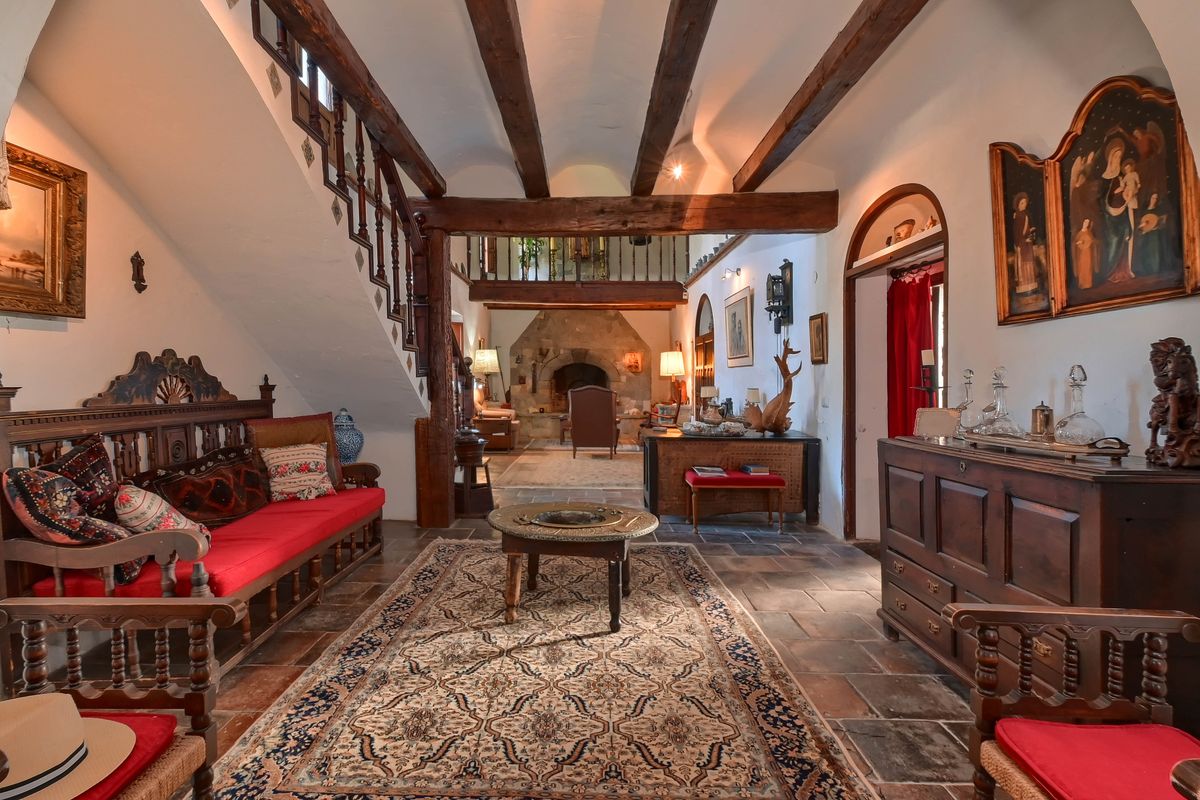 Hall traditionnel avec poutres apparentes, sol en terre cuite et mezzanine. Comprend des meubles en bois sculpté et une cheminée en pierre en arrière-plan.