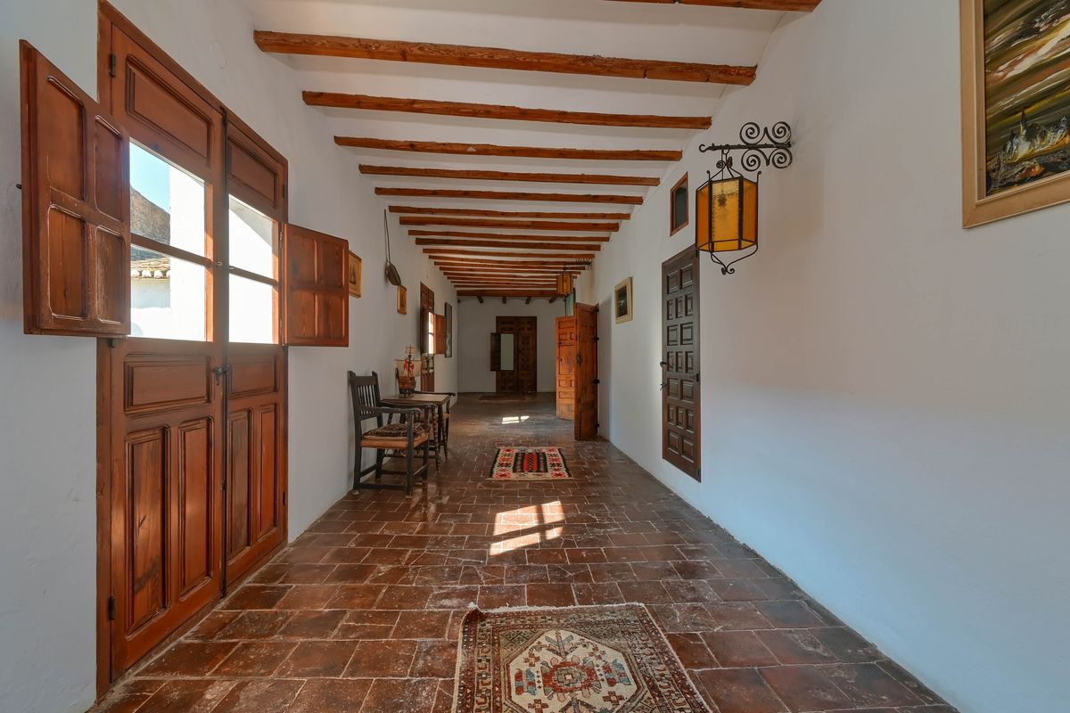 Long couloir avec poutres en bois apparentes, sol en terre cuite et portes en bois massif dotées de volets intérieurs traditionnels.