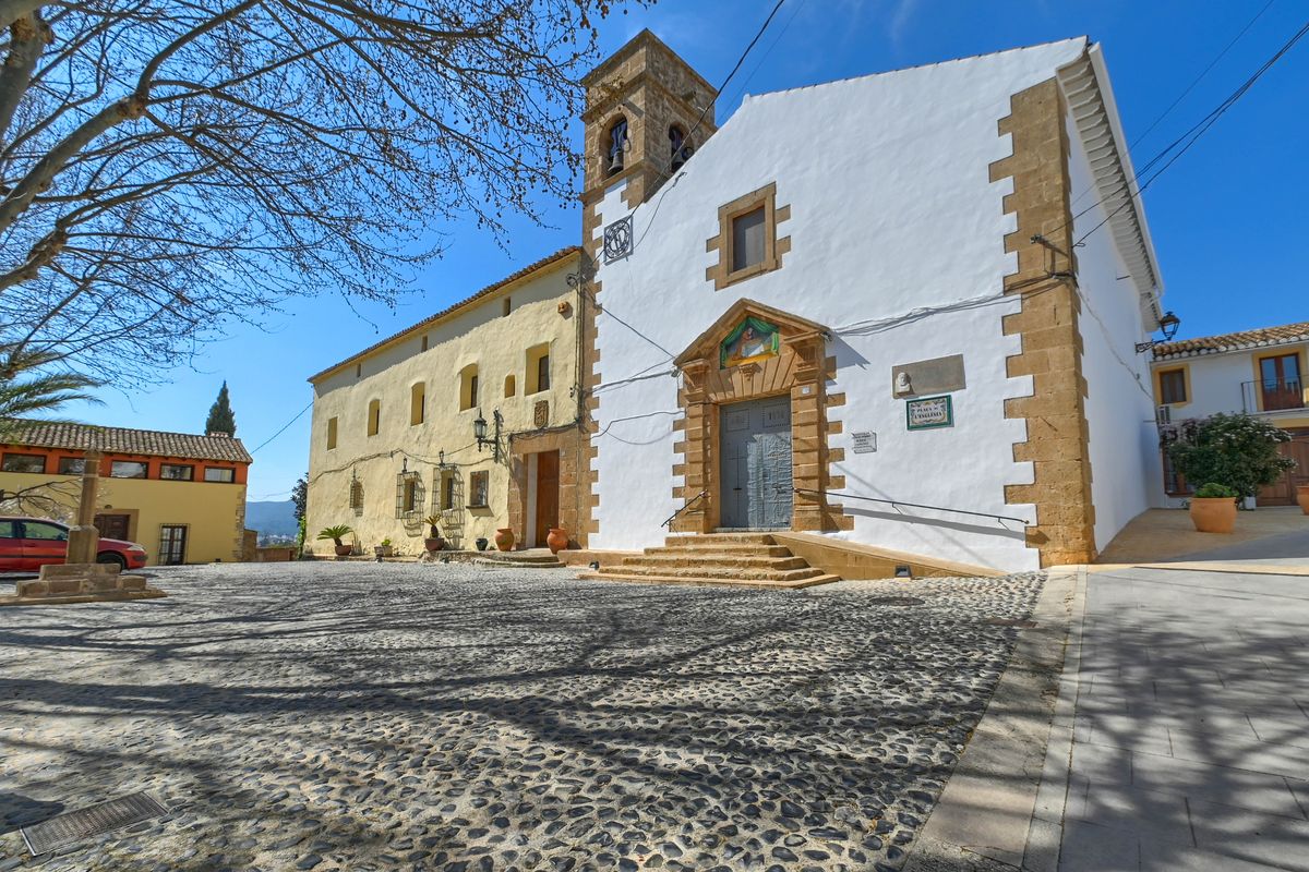 Place de l'église à Jesús Pobre avec pavés traditionnels, façade blanche aux angles en pierre de taille et clocher historique sous un ciel bleu.
