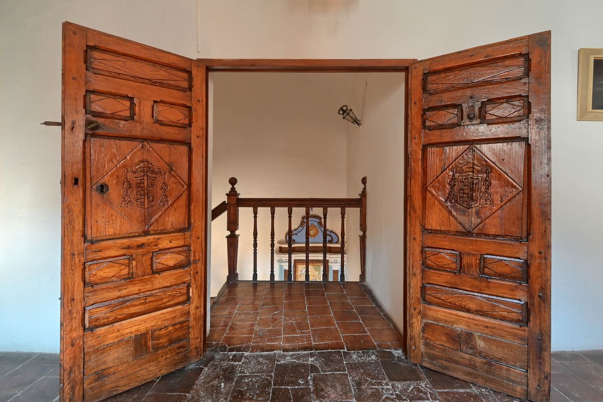 Portes doubles en bois ancien avec blasons sculptés ouvrant sur un palier en terre cuite avec balustrade d'époque et murs blancs.