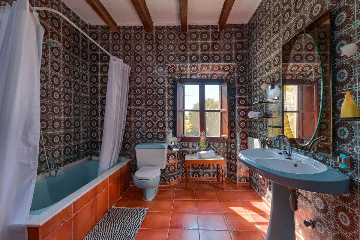 Salle de bain rustique avec carrelage géométrique, sanitaires bleu pastel, poutres en bois d'origine et sol en terre cuite naturelle.