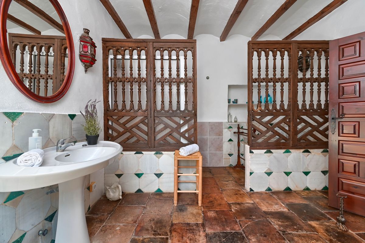 Salle de bain rustique avec poutres apparentes, sol en terre cuite, lavabo sur colonne et paravents en bois sculpté traditionnels.