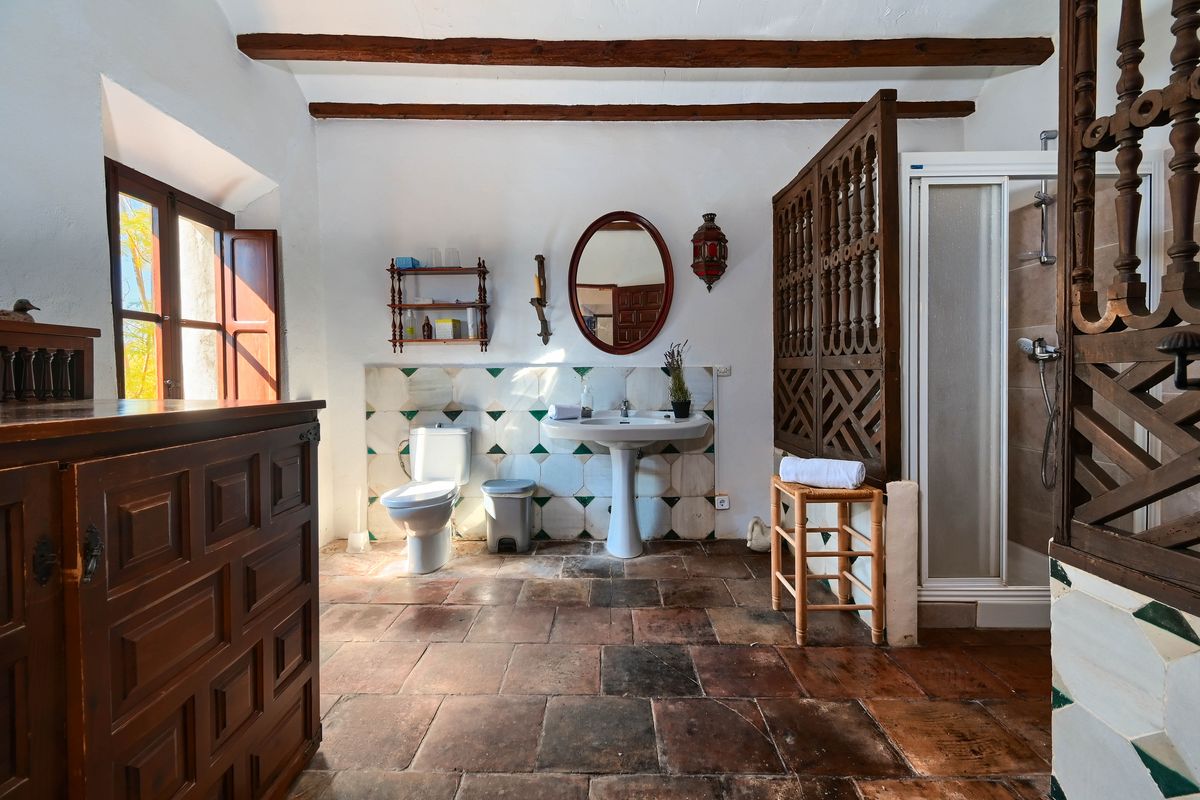 Salle de bain rustique avec sol en terre cuite, poutres apparentes et claustra en bois sculpté. Équipée d'un lavabo sur colonne et d'un carrelage mural décoratif.
