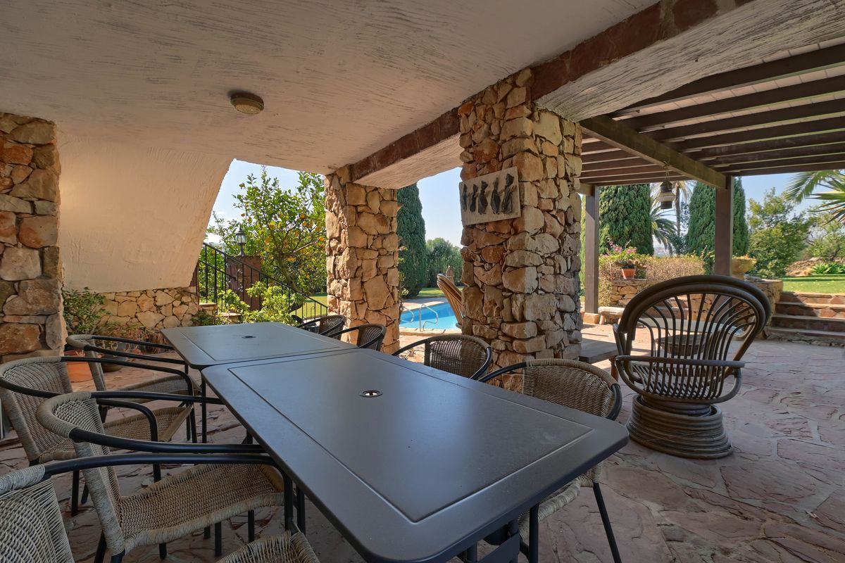 Terrasse couverte avec piliers en pierre sèche, espace dînatoire extérieur et vue sur la piscine bordée d'un jardin méditerranéen.
