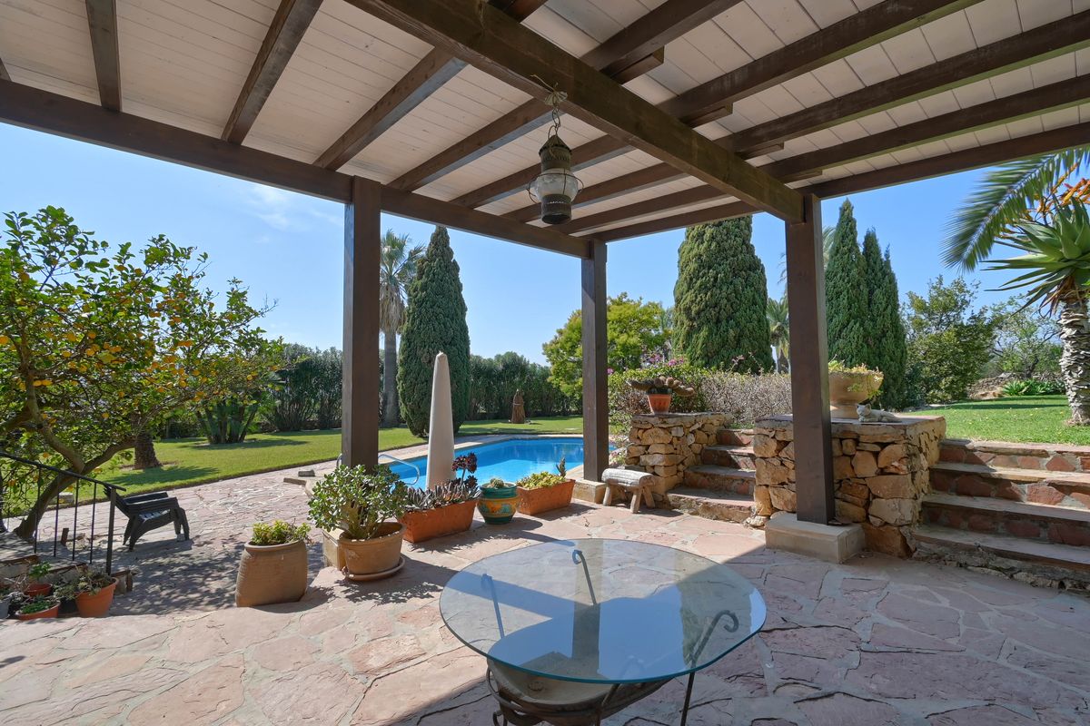 Terrasse couverte avec poutres en bois et sol en pierre, offrant une vue sur la piscine et le jardin méditerranéen arboré.