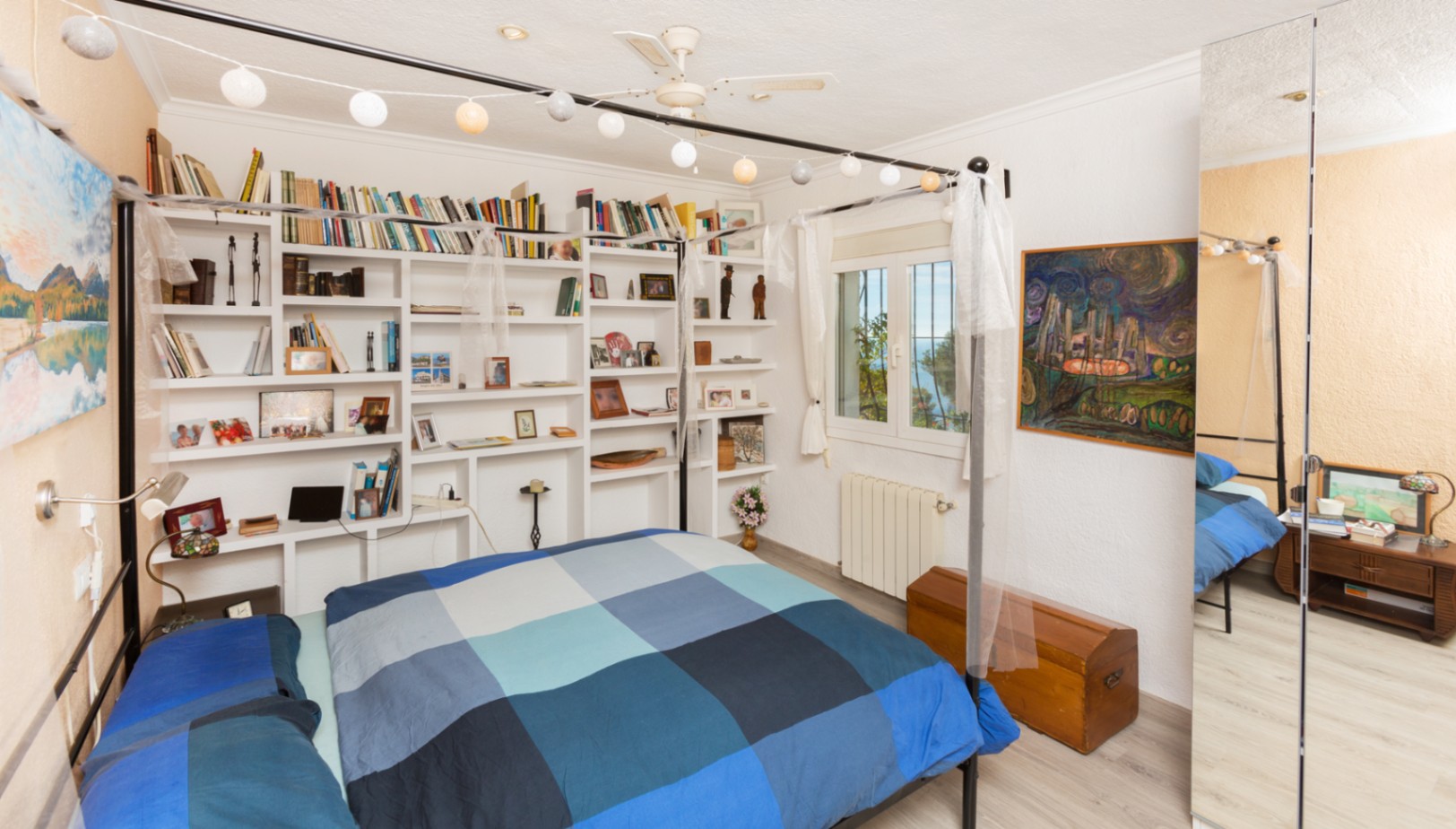 Chambre à coucher avec lit à baldaquin noir, étagères murales intégrées et grand miroir. Équipée d'un ventilateur de plafond et offrant une vue dégagée par la fenêtre.