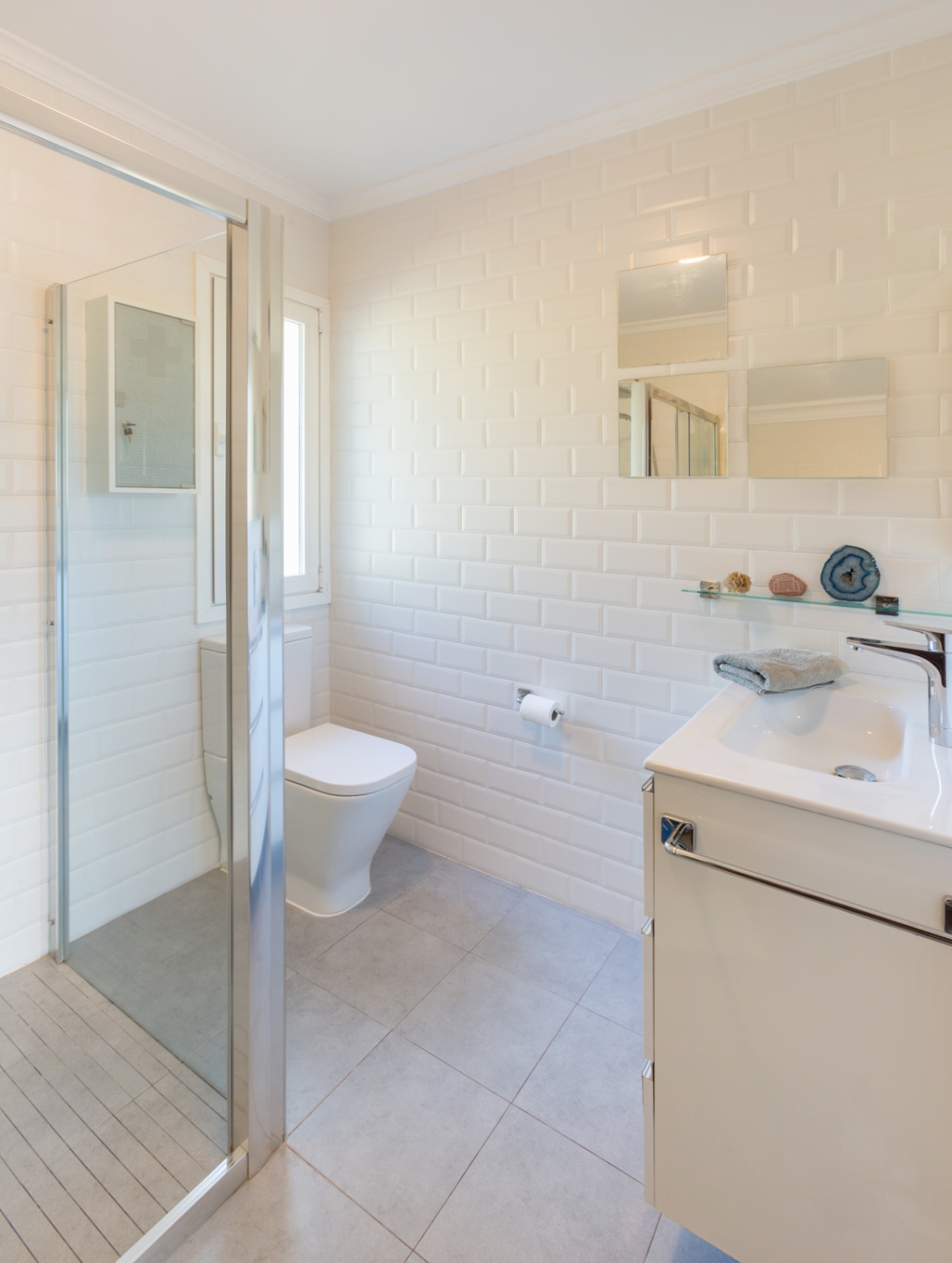 Salle de bain avec carrelage métro blanc, douche à l'italienne vitrée et meuble vasque blanc. Le sol en grès cérame gris et la fenêtre apportent clarté.