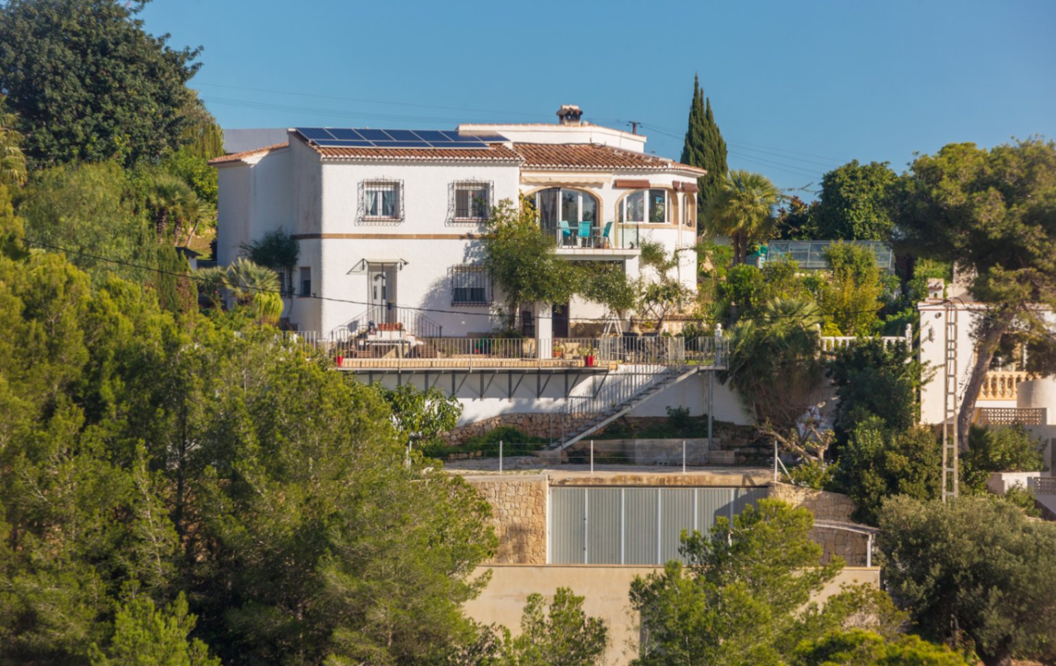 Villa méditerranéenne à plusieurs niveaux avec façades blanches, panneaux solaires et terrasses spacieuses nichées dans une végétation dense de pins.