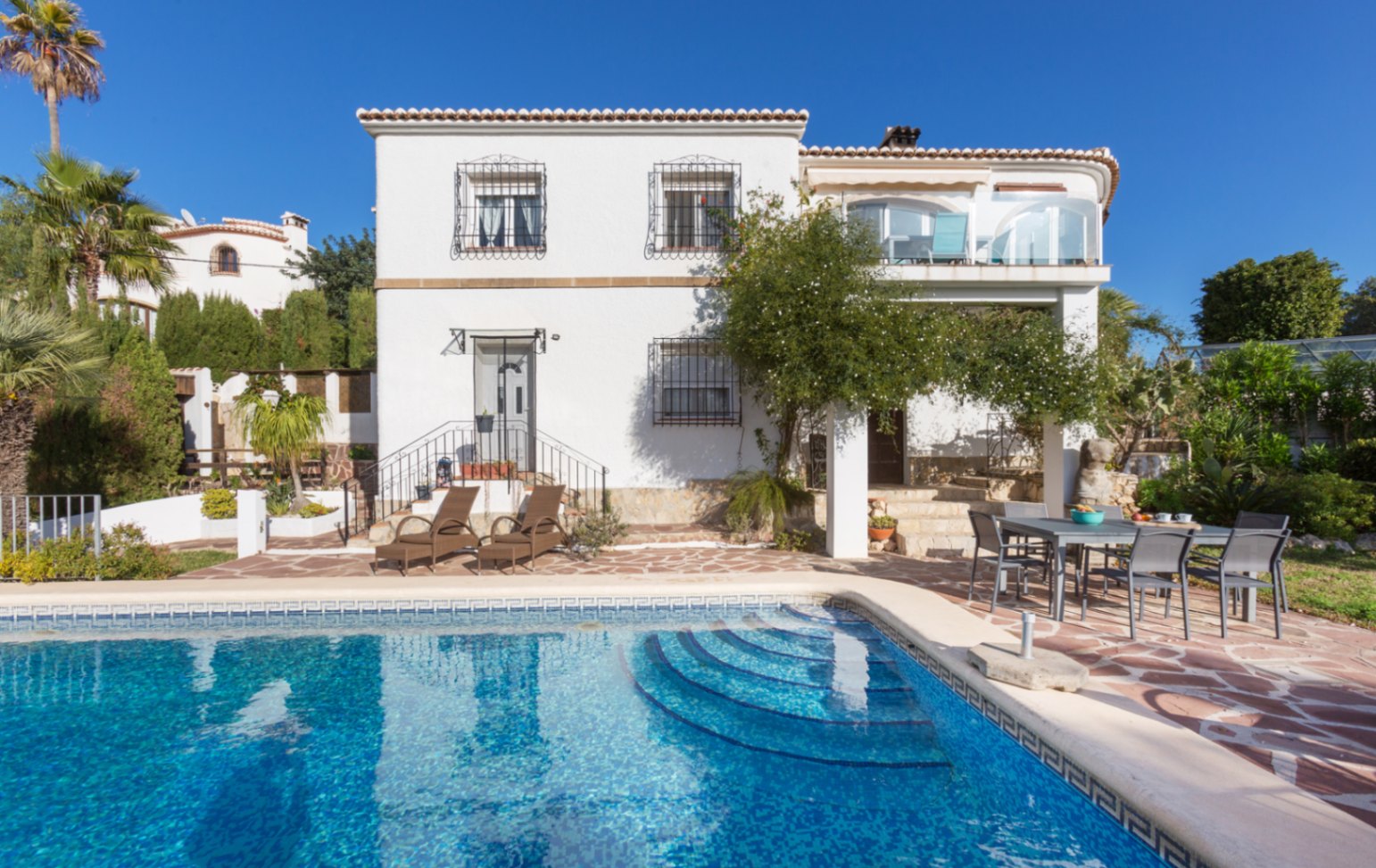 Vue extérieure de la villa avec piscine privée, terrasse en pierre naturelle, espace repas et façade blanche ornée de grilles en fer forgé.