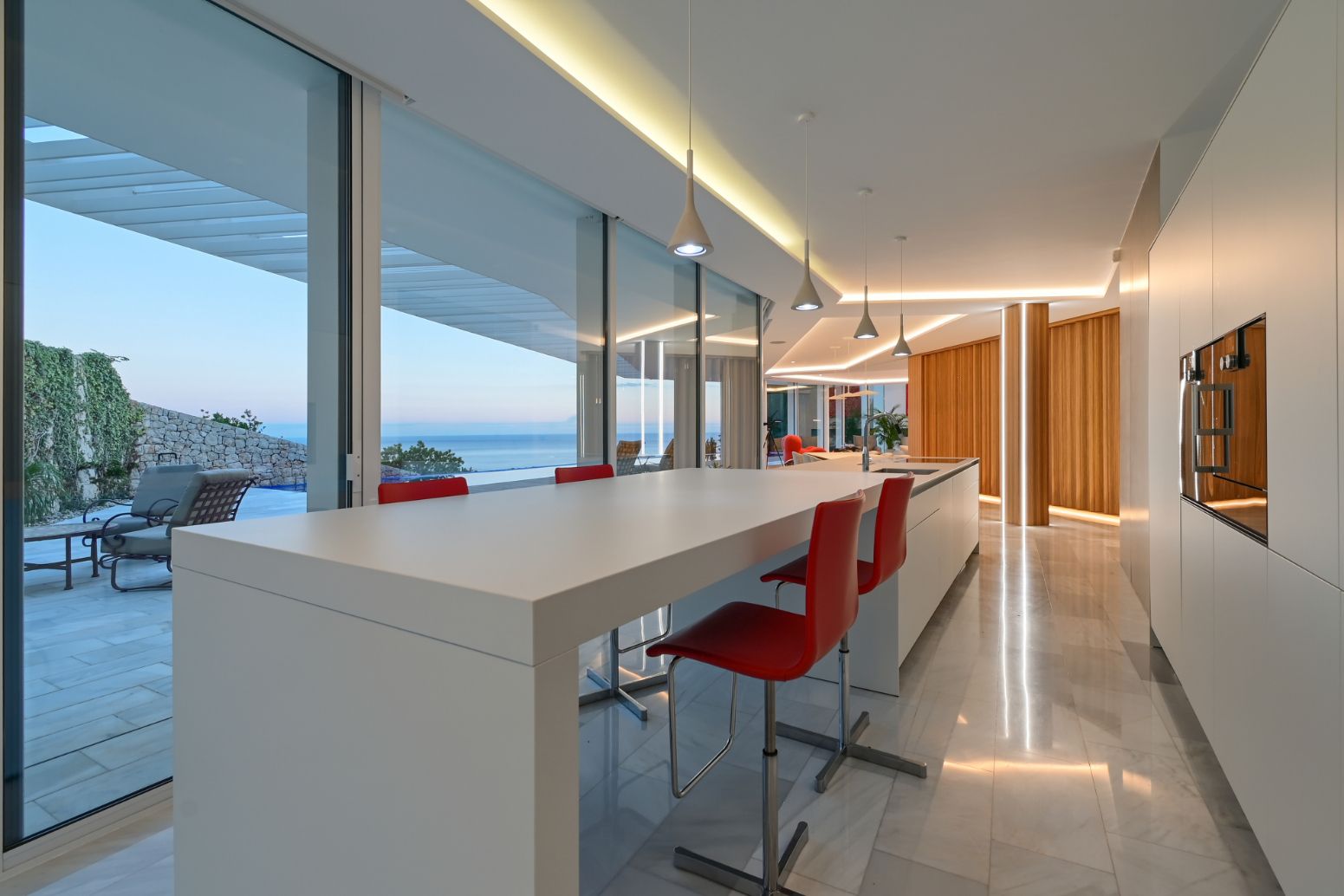Cuisine moderne avec îlot blanc minimaliste, tabourets rouges, baies vitrées offrant une vue sur la mer et éclairage LED encastré.