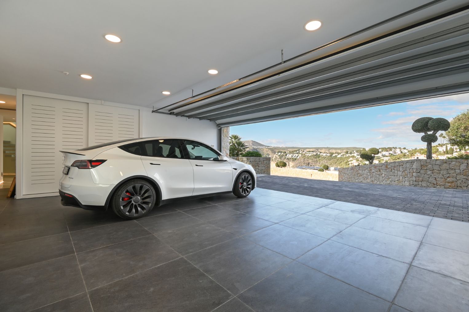 Garage moderne avec carrelage gris, spots encastrés et porte ouverte offrant une vue sur la vallée et l'allée pavée en pierre naturelle.