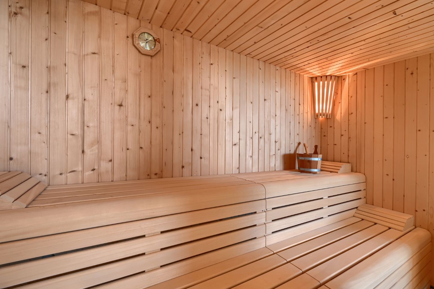 Intérieur de sauna en bois clair avec des bancs à lattes sur plusieurs niveaux, un éclairage d'angle et des accessoires en bois traditionnels.