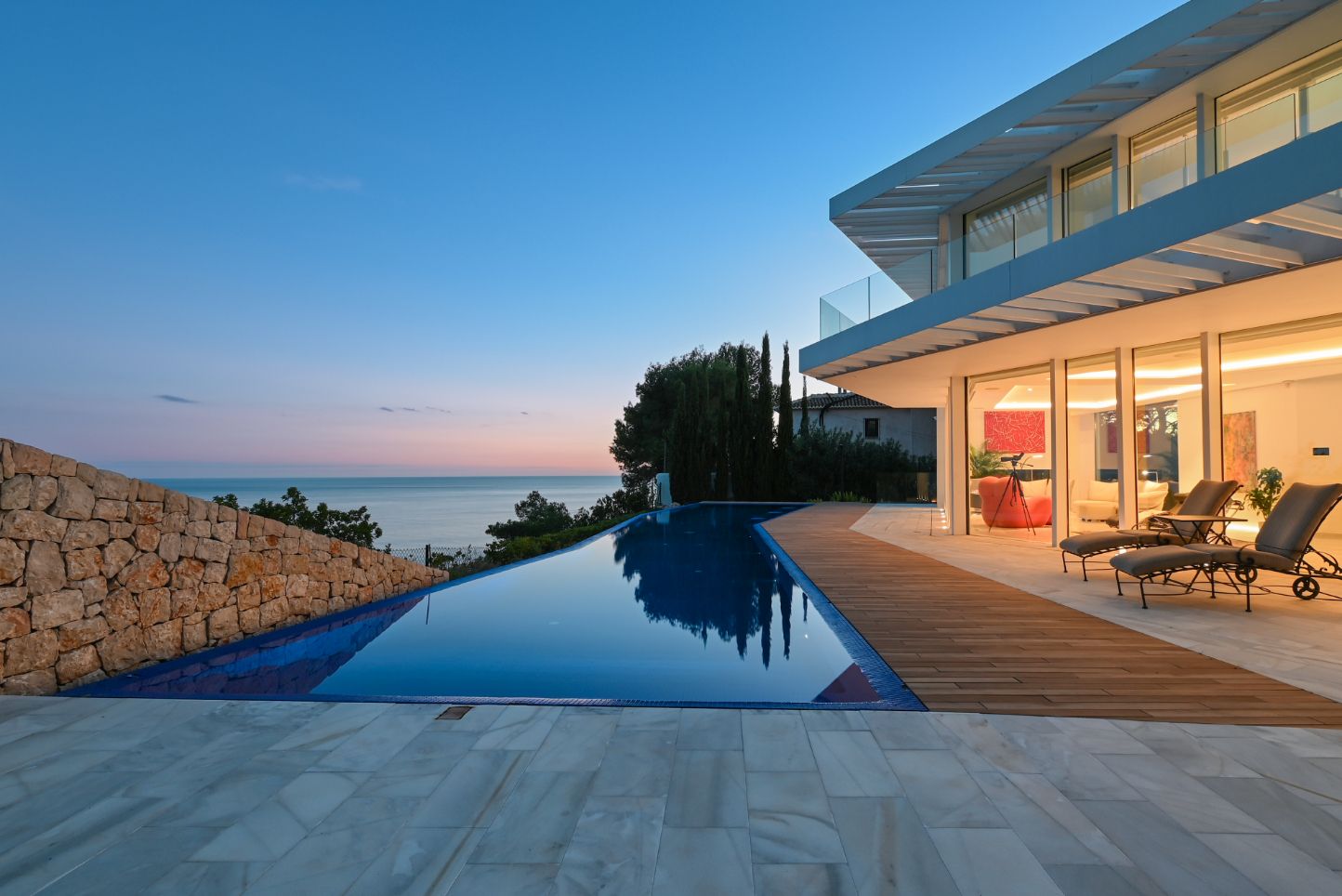 Piscine à débordement avec terrasse en bois et dallage en pierre face à la mer. Villa moderne aux larges baies vitrées et murs en pierre sèche au crépuscule.