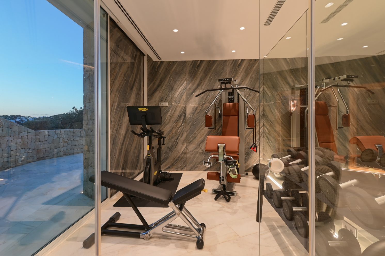 Salle de sport privée équipée de matériel Technogym, avec murs en pierre veinée et parois vitrées offrant une vue sur une terrasse extérieure en pierre.