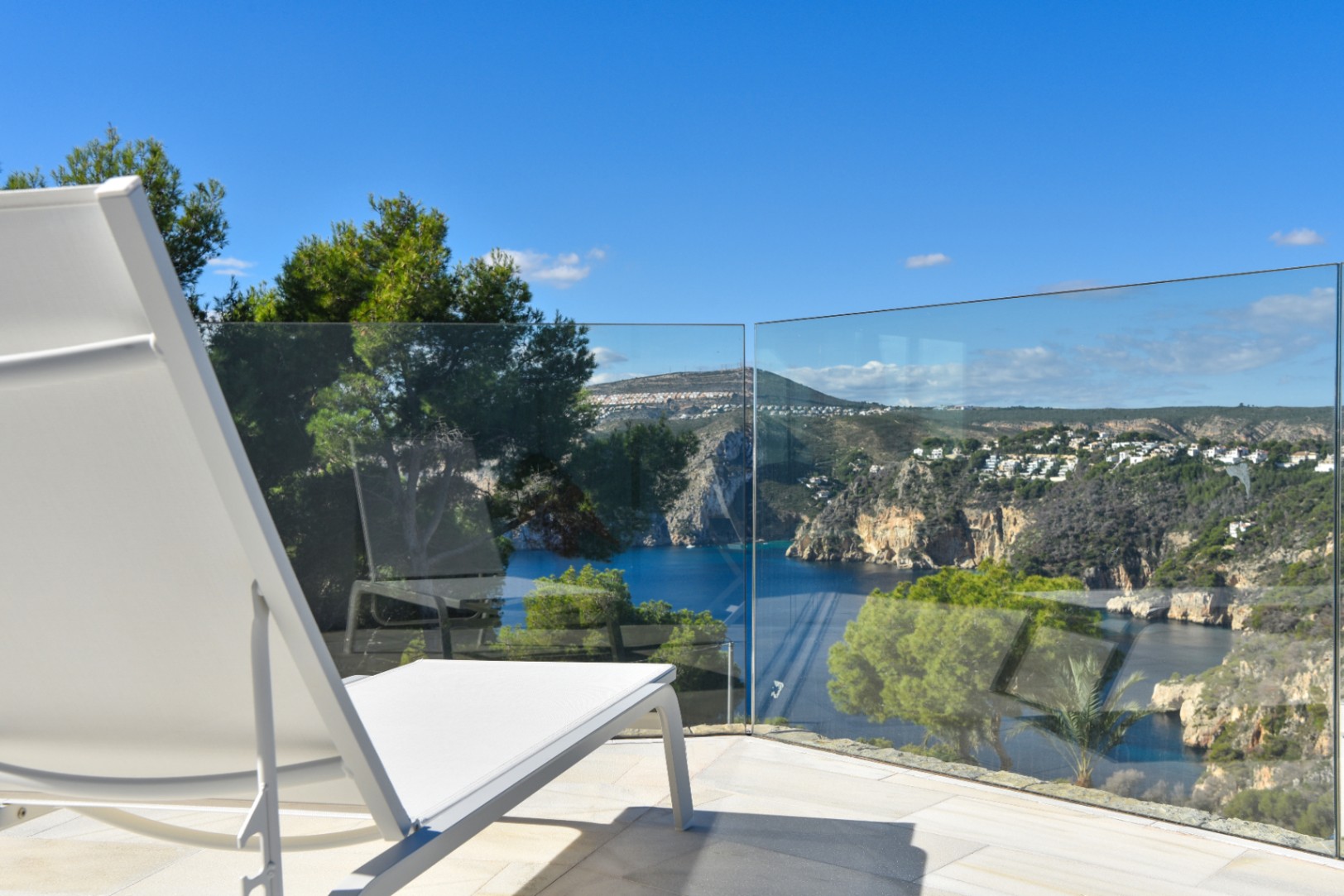 Terrasse avec chaise longue blanche et garde-corps en verre, offrant une vue dégagée sur une baie méditerranéenne et des falaises escarpées.