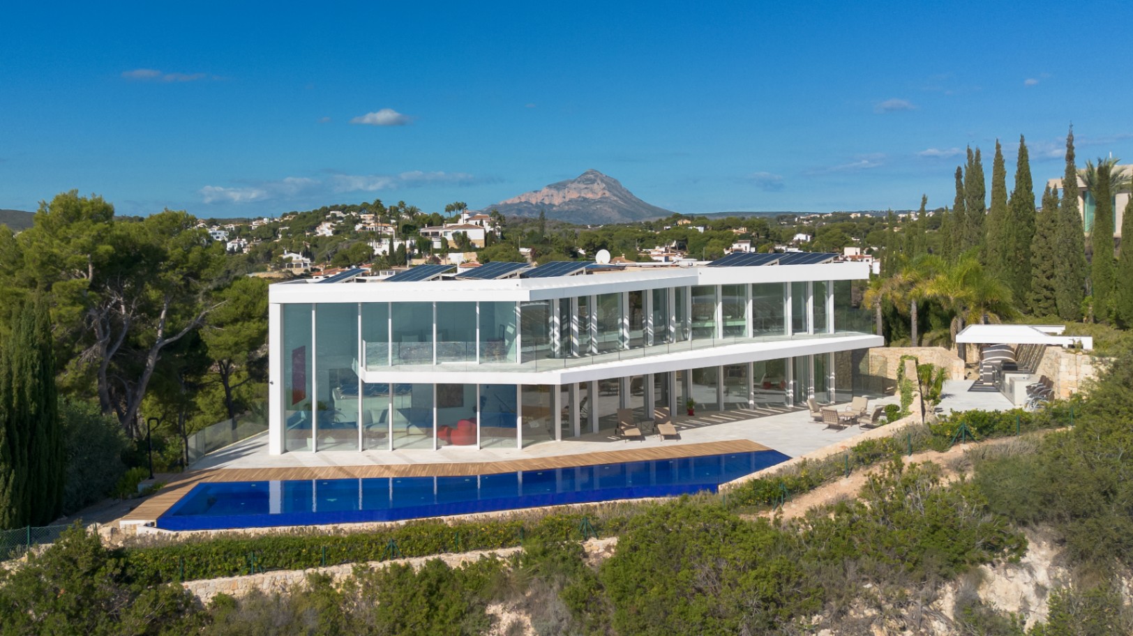 Villa contemporaine à deux étages avec façades vitrées, piscine à débordement, panneaux solaires et vue panoramique sur la montagne Montgó.