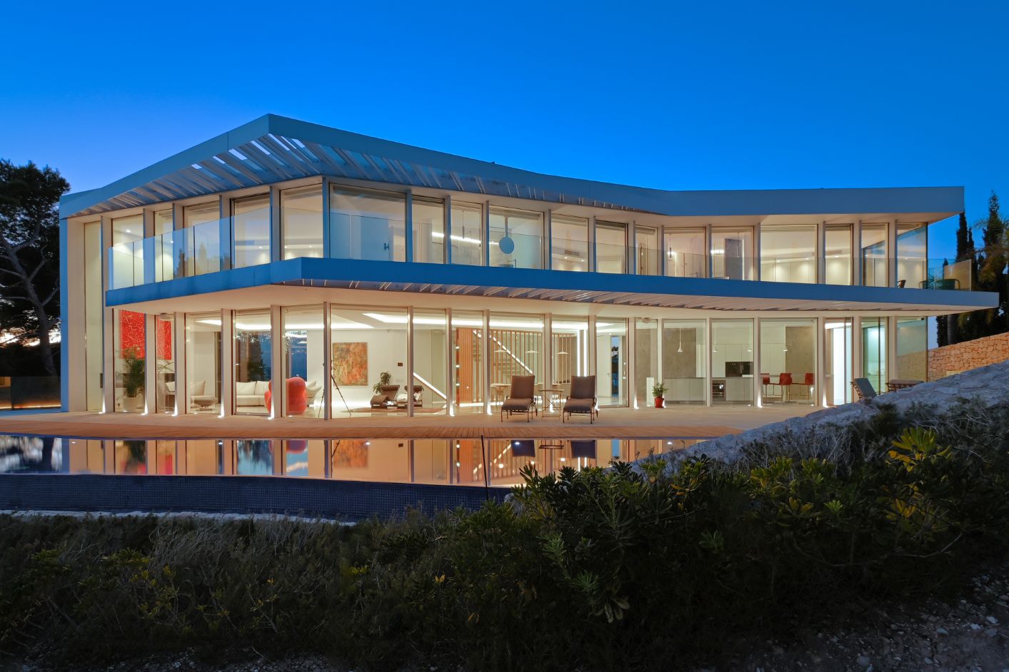 Villa moderne sur deux niveaux avec façades vitrées, vaste terrasse avec piscine à débordement et éclairage intérieur visible au crépuscule.