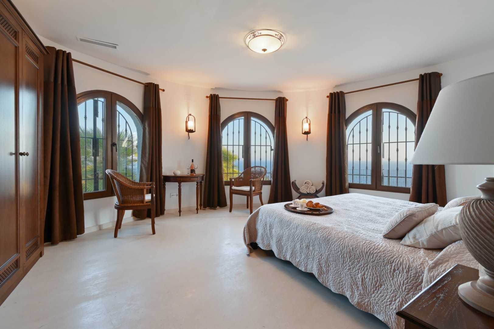 Chambre spacieuse avec mur incurvé, trois fenêtres en plein cintre, menuiseries en bois sombre et vue dégagée sur la mer Méditerranée.