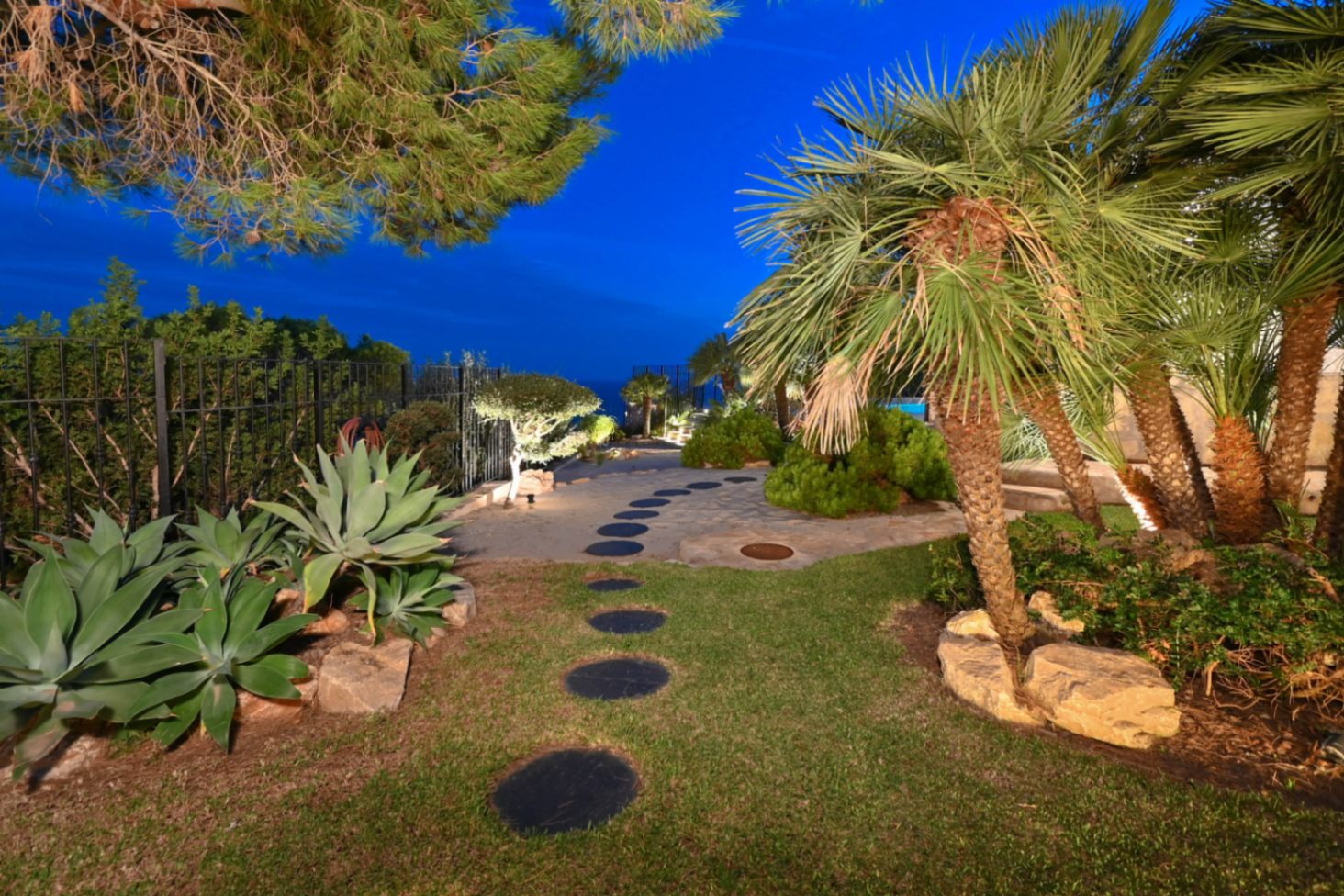 Chemin de dalles circulaires dans un jardin méditerranéen avec palmiers, agaves et éclairage nocturne intégré sous un ciel de crépuscule.