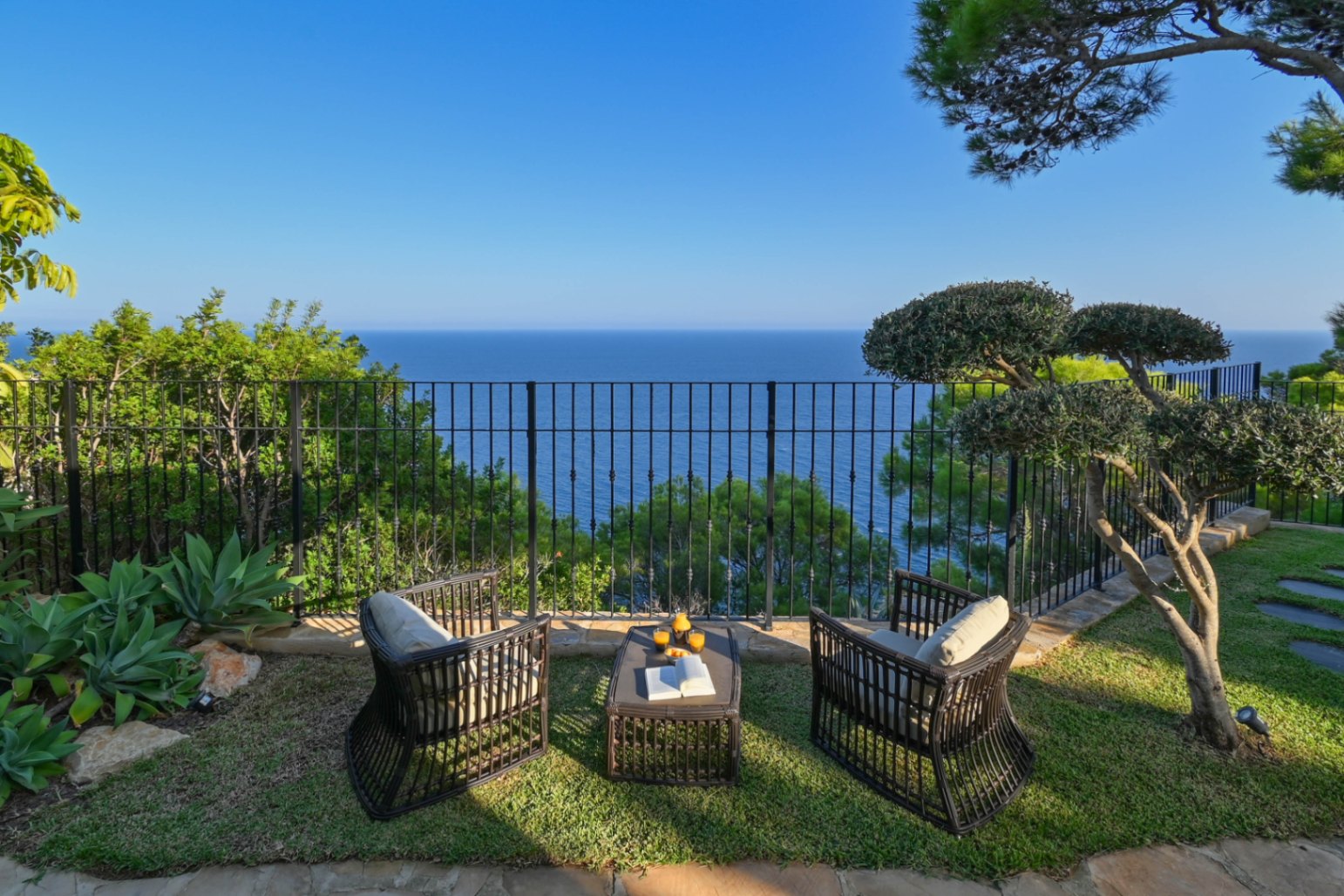 Coin salon avec fauteuils en osier sombre et table sur pelouse, offrant une vue panoramique sur la mer derrière une balustrade en fer forgé.