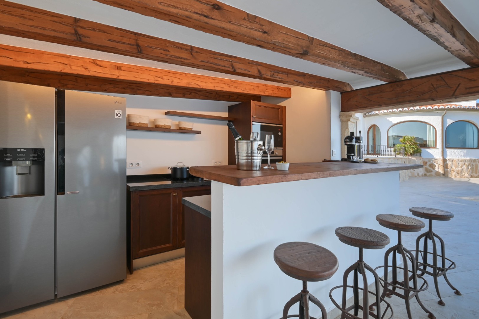 Cuisine rustique avec poutres en bois apparentes, réfrigérateur en inox et bar blanc. Des tabourets de style industriel complètent cet espace ouvert sur la terrasse en pierre.