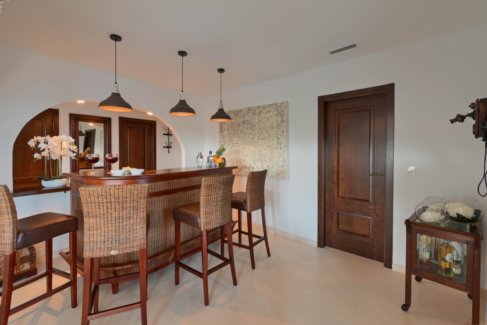 Espace bar intérieur avec comptoir en bois courbé et rotin, tabourets hauts, suspensions industrielles et portes en bois massif sur fond de murs blancs épurés.