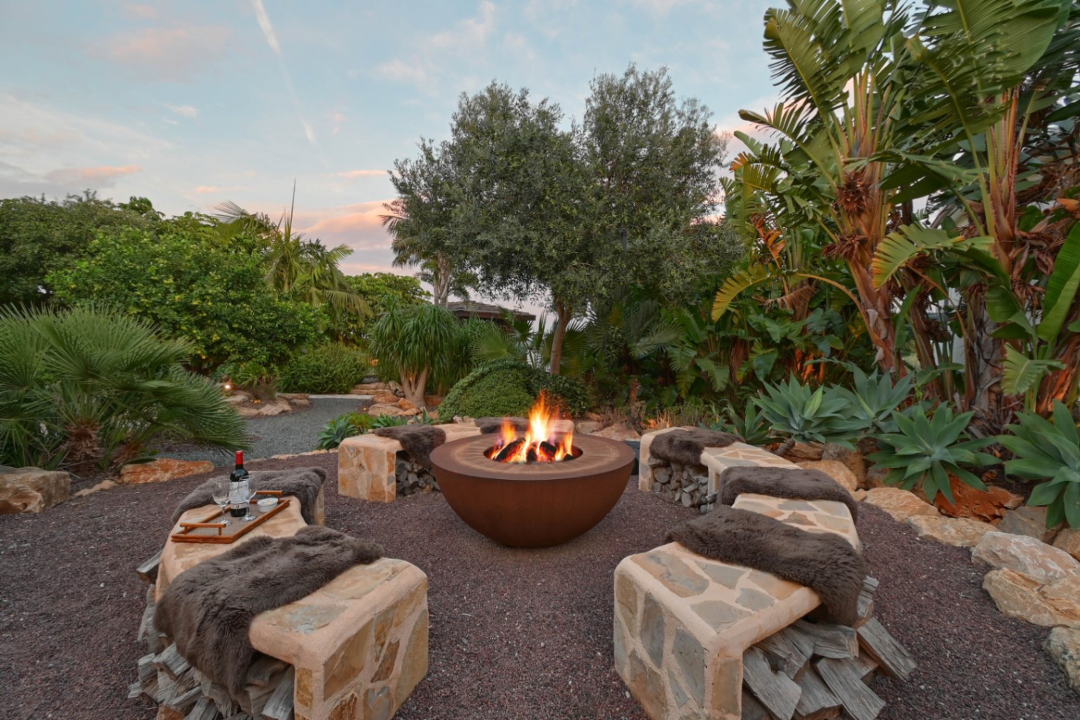 Espace brasero circulaire en acier Corten avec bancs en pierre, rangement pour bois intégré et jardin tropical luxuriant sous un ciel de crépuscule.