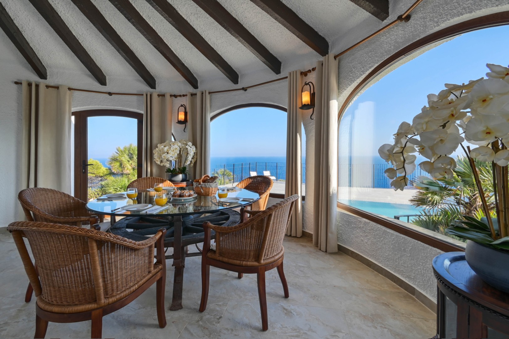 Espace repas circulaire avec table en verre et chaises en osier, caractérisé par son plafond à poutres sombres et ses ouvertures voûtées offrant une vue sur la mer.