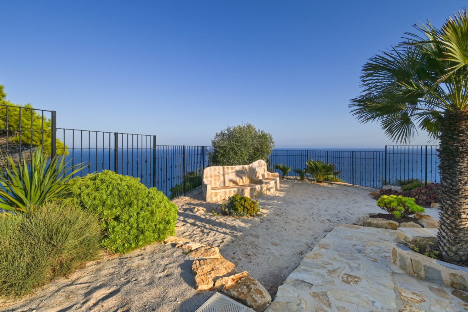 Point de vue extérieur avec banc en pierre, clôture en fer noir et vue panoramique sur la mer Méditerranée, agrémenté de palmiers et d'arbustes.