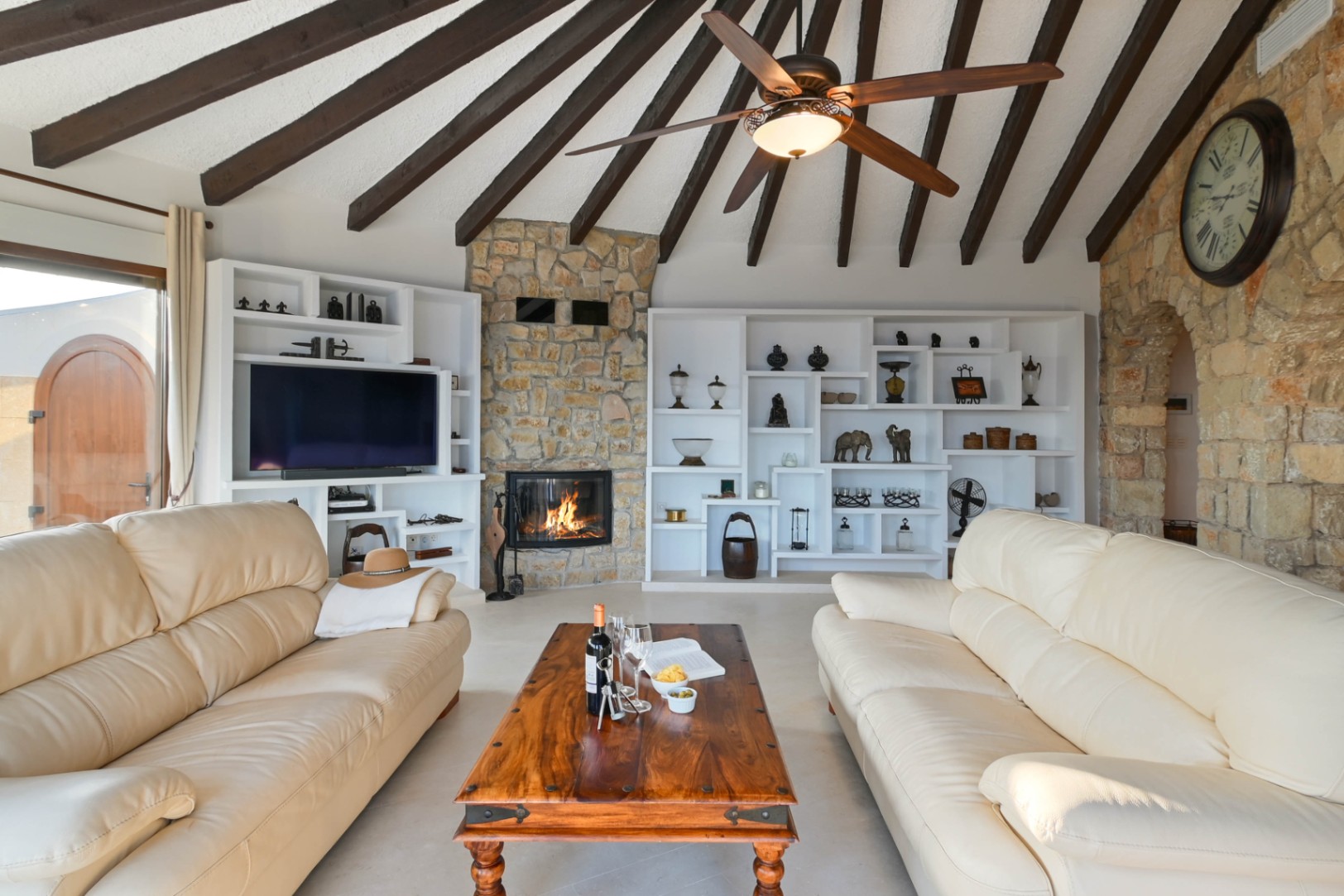 Salon avec plafond à poutres apparentes, cheminée en pierre et étagères blanches intégrées. Équipé de canapés en cuir crème et d'une table basse en bois massif.