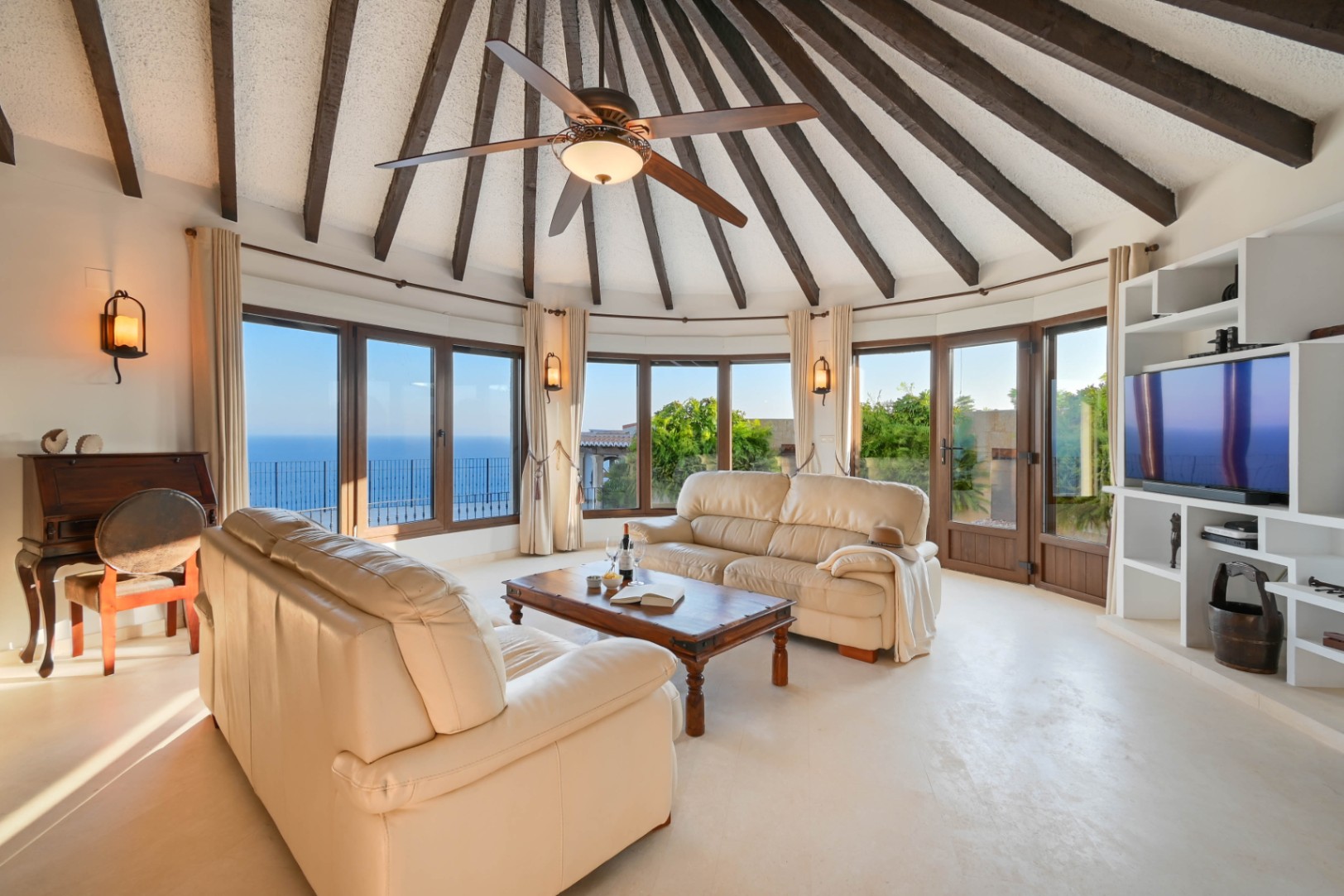 Salon circulaire doté d'un plafond voûté aux poutres apparentes, de canapés en cuir et de grandes fenêtres offrant une vue panoramique sur la mer.