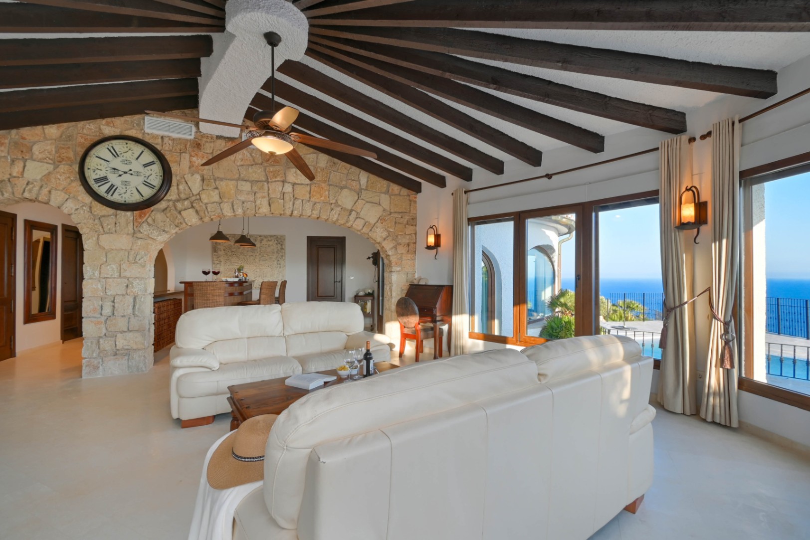 Salon spacieux avec plafond voûté à poutres de bois radiales, arche en pierre naturelle et grandes baies vitrées offrant une vue panoramique sur la mer.