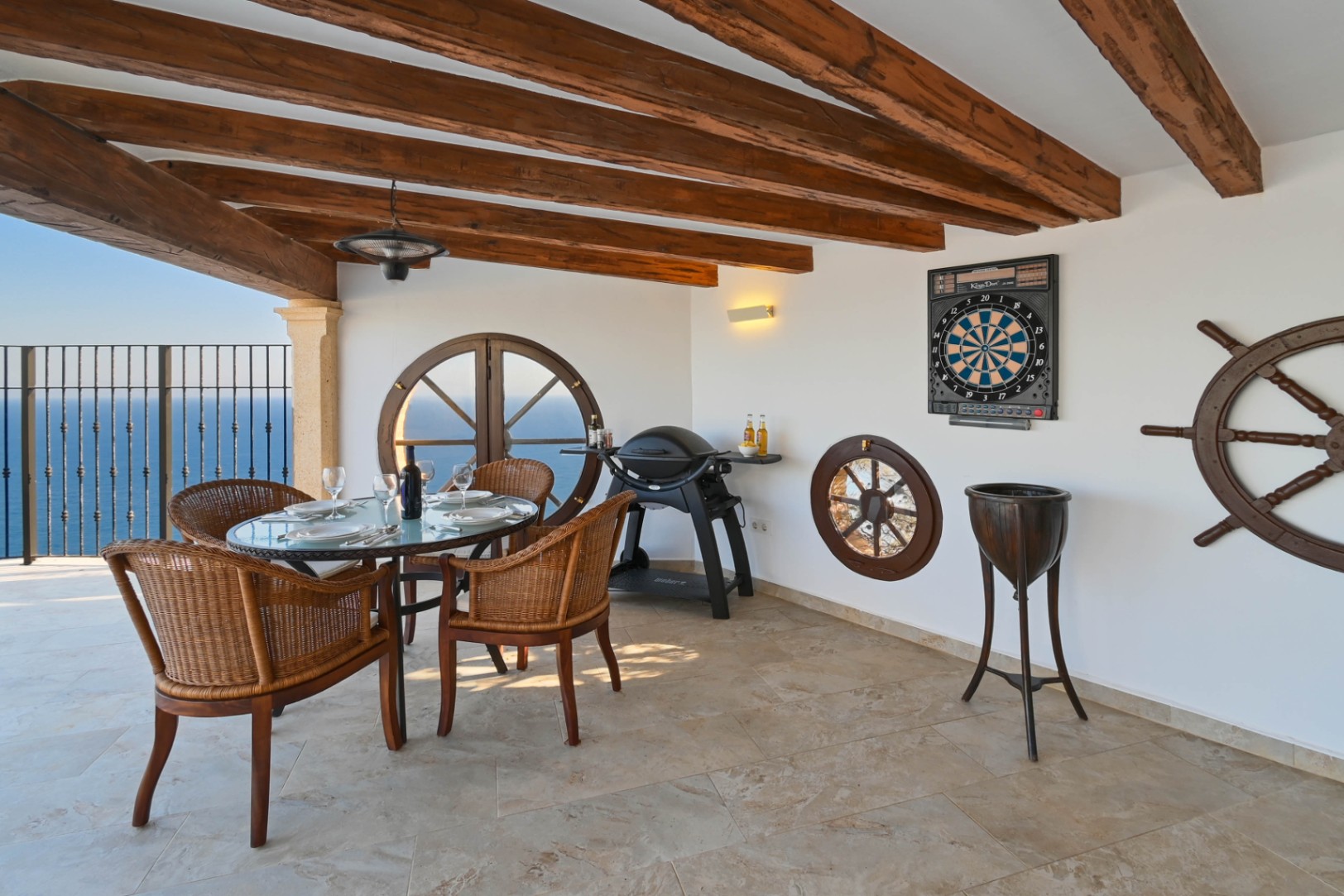 Terrasse couverte avec poutres apparentes, fenêtres œil-de-bœuf et vue mer. Comprend un ensemble repas en osier, un barbecue à gaz et une décoration thématique marine.