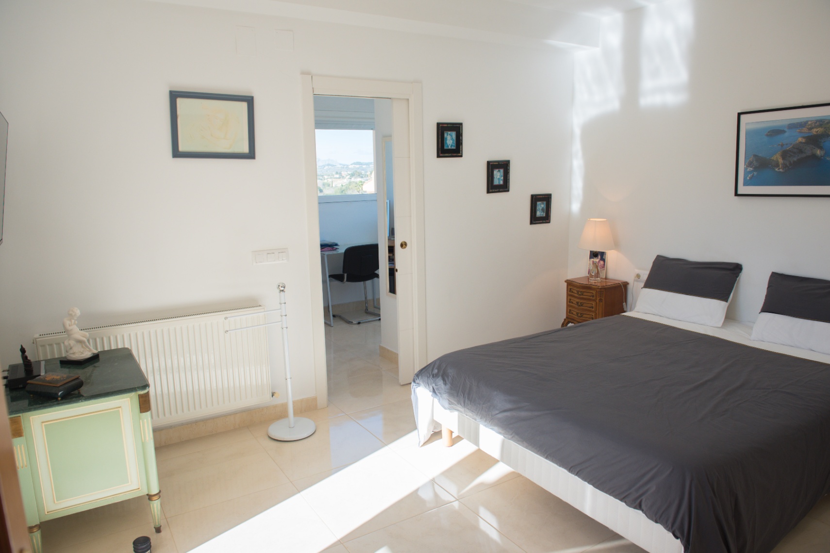 Chambre à coucher avec carrelage clair, lit double et mobilier vintage. Une porte ouverte mène à un bureau privé lumineux offrant une vue dégagée.