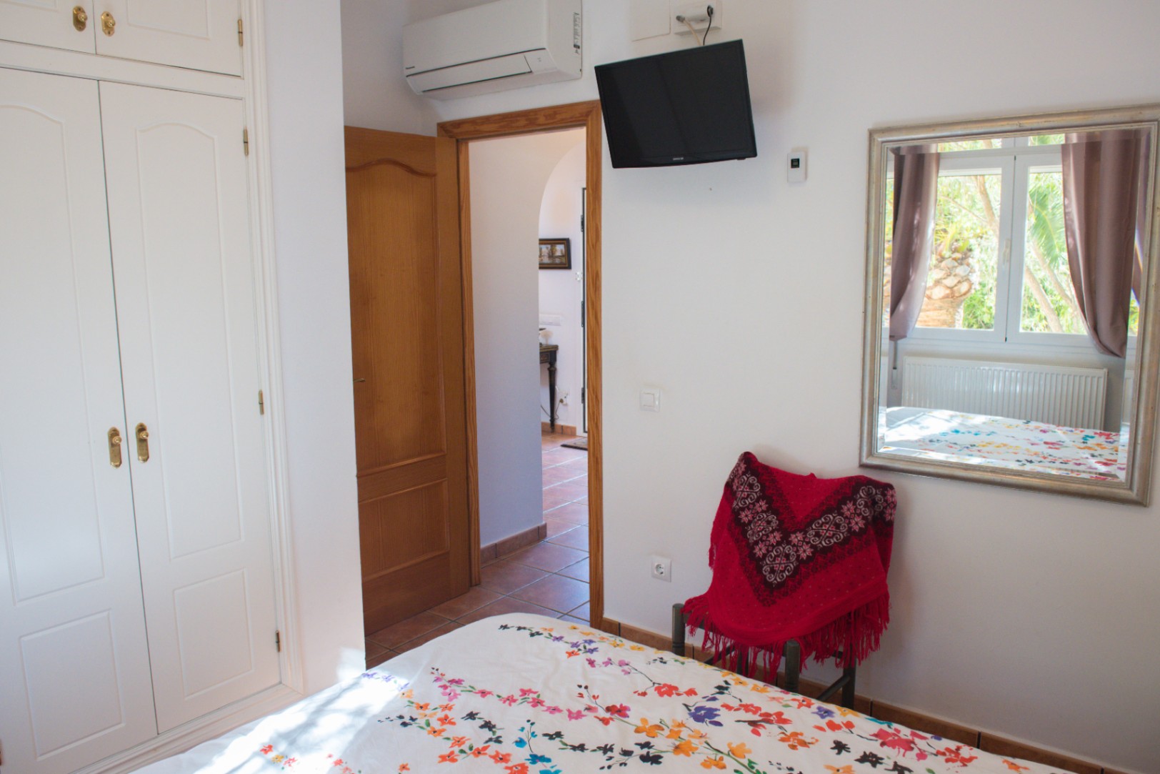 Chambre avec armoire encastrée blanche, télévision murale et climatisation. Un miroir reflète la fenêtre et une porte en bois mène au couloir carrelé.
