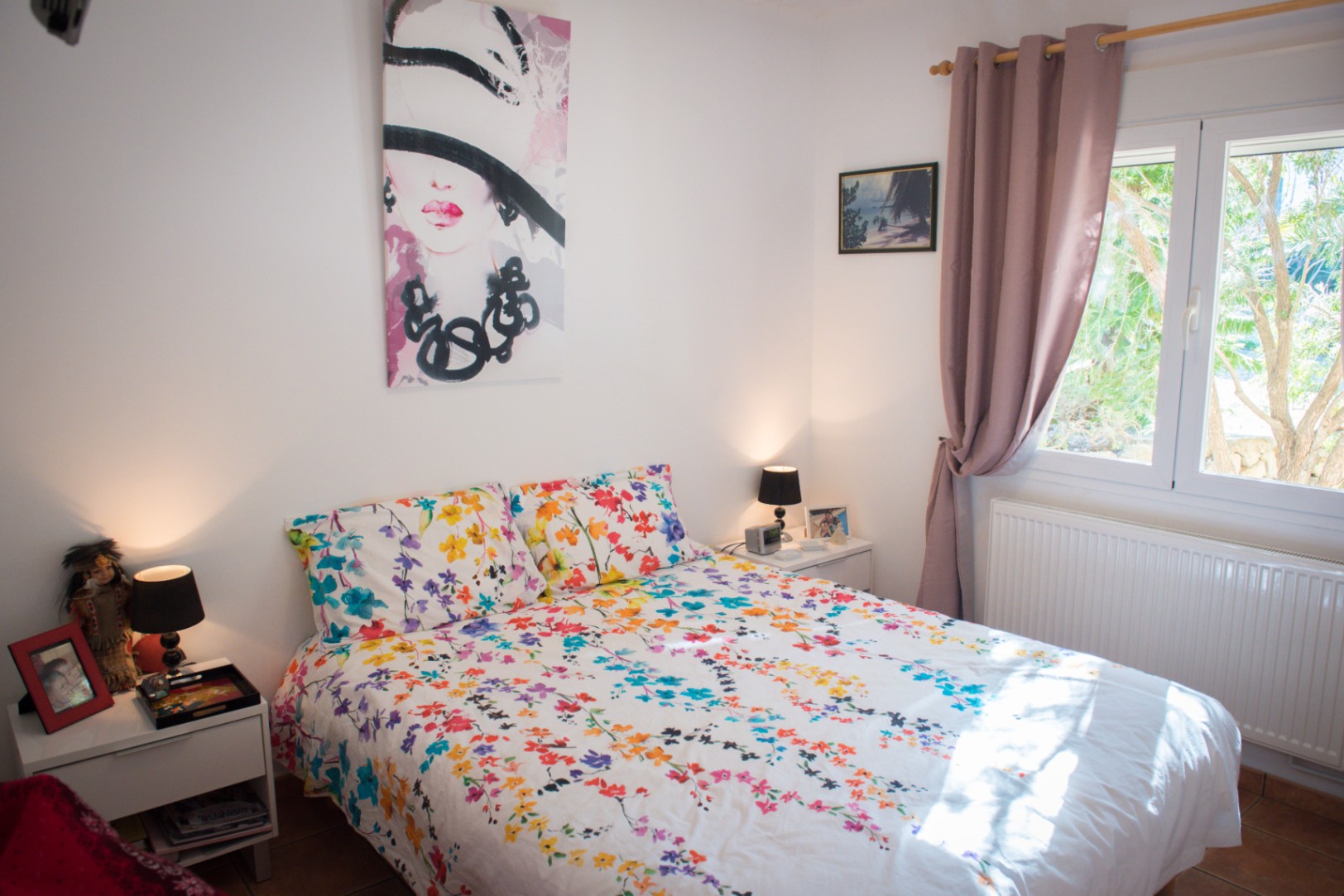 Chambre lumineuse avec parure de lit fleurie, grand tableau d'art moderne, tables de chevet blanches et fenêtre avec rideaux mauves offrant une vue sur le jardin.