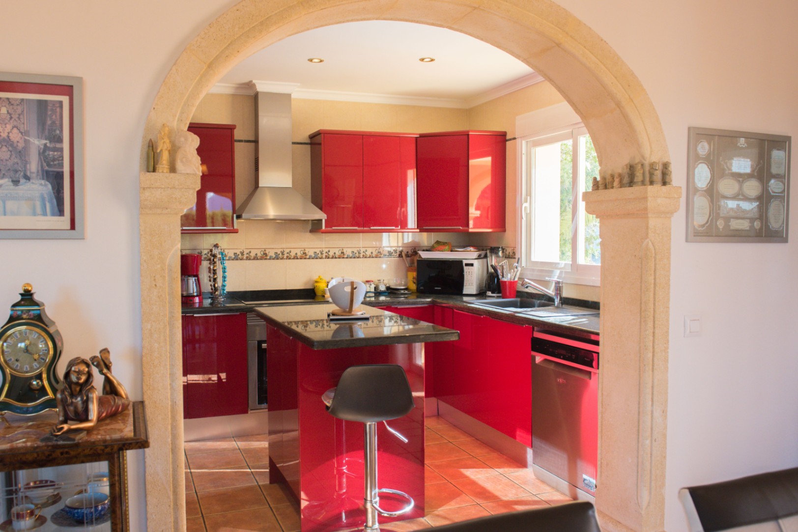 Cuisine aux meubles rouges brillants et plans de travail en granit, vue à travers une arche en pierre. Équipée d'un îlot central et d'appareils en inox.